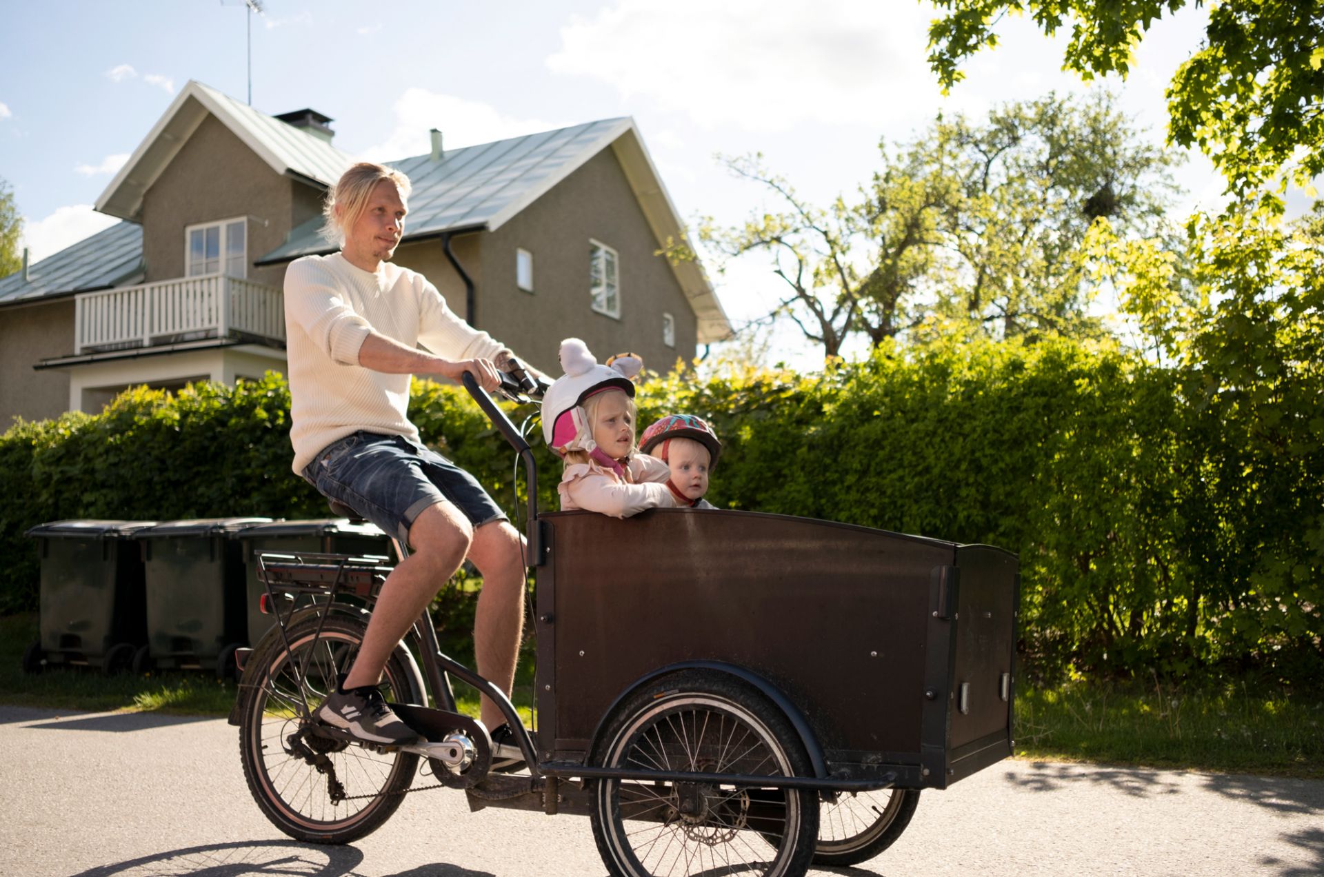 En man cyklar på en lastcykel där två barn sitter på flaket framför cykeln. De cyklar framför en grön häck.